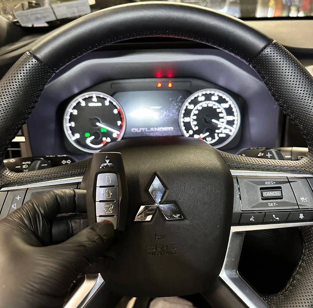 Mitsubishi Outlander smart key held in front of the steering wheel with the Mitsubishi three-diamond emblem and OUTLANDER badge visible on the dashboard